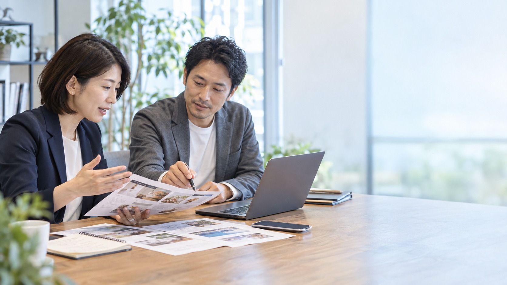 2人が机に並び、印刷したLPの見本とノートパソコンを見ながら、広告とLPの見直しを相談している様子